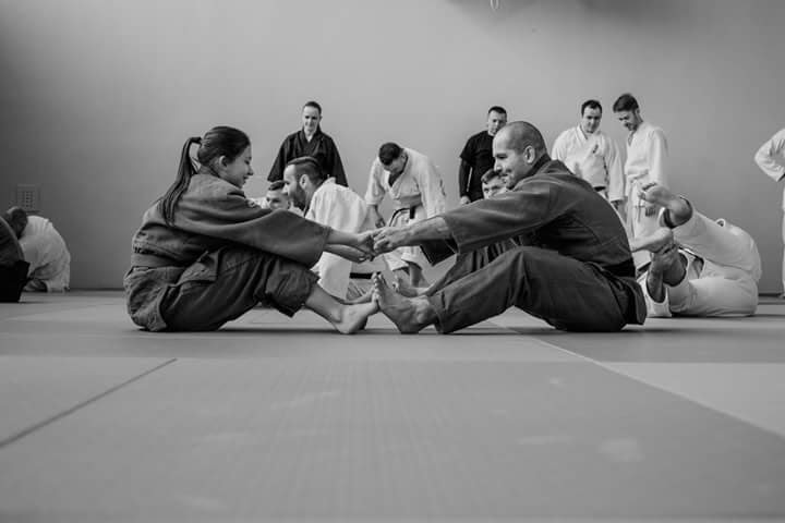 Kobieta i mężczyzna w judogach siedzą na macie, stykają sie stopami i trzymają się za ręce. W tle inni judocy.