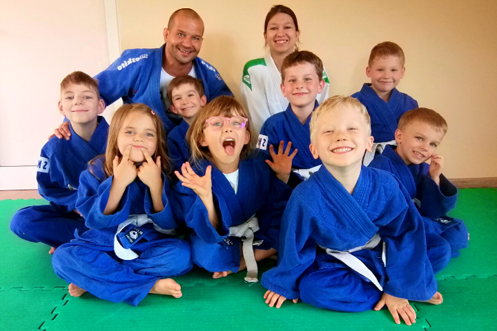 Grupka uśmiechniętych dzieci w niebieskich judogach i dwoje trenerów mężczyzna i kpbieta, siedzą na zielonaj macie.